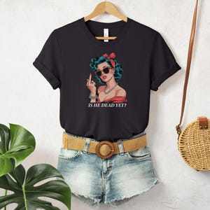 May include: Black t-shirt with a graphic of a woman with red hair and sunglasses, giving a middle finger. The text "Is he dead yet?" is printed below the graphic.