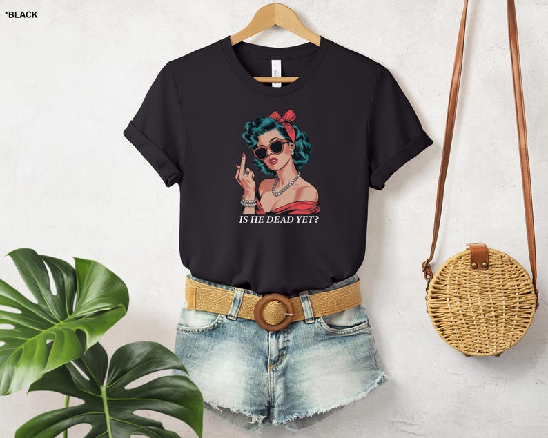 May include: Black t-shirt with a graphic of a woman with red hair and sunglasses, giving a middle finger. The text "Is he dead yet?" is printed below the graphic.
