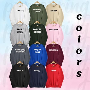 May include: A collection of 12 sweatshirts in various colors, including white, military green, sand, sport grey, forest green, soft pink, dark grey heather, royal blue, maroon, black, navy, and red. Each sweatshirt has a different color name printed on the front.