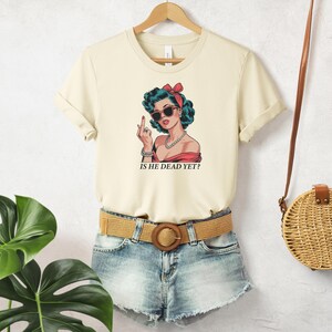 May include: A cream colored t-shirt with a cartoon image of a woman with blue hair and sunglasses. The woman is wearing a red and white dress and has a pearl necklace. The text "IS HE DEAD YET?" is printed below the image.
