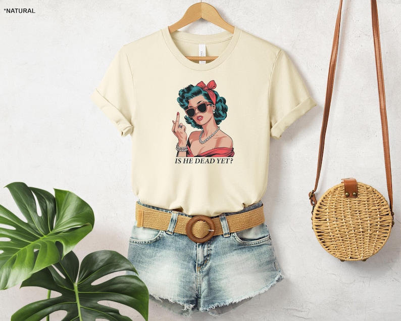 May include: A cream colored t-shirt with a cartoon image of a woman with blue hair and sunglasses. The woman is wearing a red and white dress and has a pearl necklace. The text "IS HE DEAD YET?" is printed below the image.