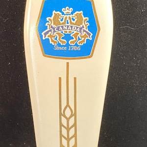May include: A white Molson Golden beer tap handle with gold accents. The handle features the Molson Golden logo, a blue and gold emblem with the word "CANADA" and the year "Since 1786". A gold wheat stalk design is also present.