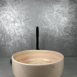 Puede incluir: Un lavabo redondo de cerámica color melocotón claro con un grifo negro. El lavabo tiene un acabado liso y brillante y un sutil patrón de vetas de madera natural. El grifo es de diseño sencillo y moderno.