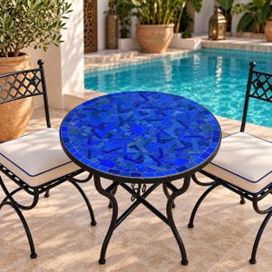 May include: A round bistro table featuring a vibrant blue mosaic tile top with an intricate geometric pattern, supported by a black metal frame, flanked by two matching black metal chairs with off-white cushions trimmed in blue piping.