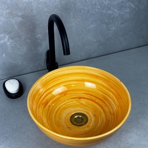 Puede incluir: Un lavabo redondo de cerámica amarilla y naranja con un diseño en espiral. El lavabo tiene un desagüe de latón y un grifo negro. A la izquierda hay un dispensador de jabón blanco y negro.