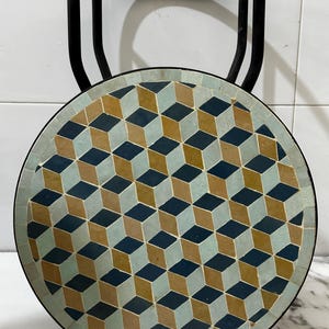 May include: A round side table with a black metal frame and a mosaic top. The mosaic design features a repeating pattern of blue, tan, and teal cubes. The table is approximately 24 inches in diameter.