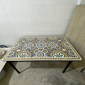 May include: Rectangular mosaic tile table with a black metal frame. The tabletop features a colorful geometric pattern with yellow, blue, green, and brown tiles. The table is approximately 48 inches long and 24 inches wide.