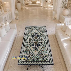 Puede incluir: Mesa de centro rectangular con marco negro y diseño con incrustaciones ornamentadas. La superficie presenta un fondo blanco con un patrón simétrico de elementos florales y geométricos verdes, azules y dorados. La mesa está sostenida por patas de metal negras y curvas.