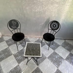May include: Two black wrought iron chairs with round black velvet seats and decorative backrests, flanking a small square table. The table has a black and white tiled top and black metal legs. The floor is tiled in a black and white checkered pattern.