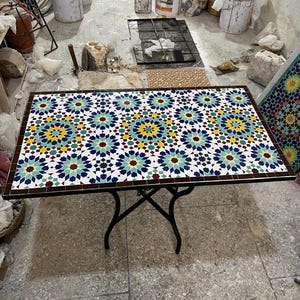Peut inclure: Une table rectangulaire dotée d'un plateau en mosaïque avec un motif géométrique en étoile bleu, jaune, vert et blanc, encadré par une bordure marron foncé. La table repose sur une base en métal noir avec quatre pieds incurvés qui se croisent.
