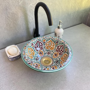 May include: A decorative ceramic sink with a colorful floral pattern in shades of teal, orange, and burgundy. The sink has a gold-colored drain and is paired with a black faucet, a soap dish, and a floral-patterned soap dispenser.