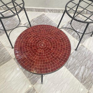 May include: A round table with a mosaic tile top in dark red. The table has three thin, black metal legs. Two matching plant stands with black metal frames are in the background.