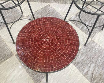 May include: A round table with a dark red mosaic tile top featuring a circular pattern and a black metal frame, accompanied by two matching black metal stools with circular grid seats, all arranged on a light gray tiled floor surface.