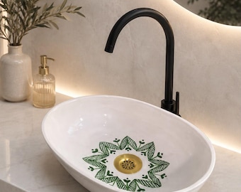 May include: An oval white ceramic vessel sink featuring a green geometric floral pattern painted at the center around a gold-toned drain. A tall matte black gooseneck faucet is mounted behind the basin, which sits on a light-colored stone countertop.