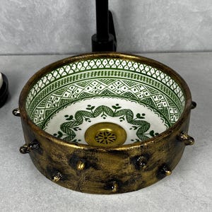 Puede incluir: Un lavabo decorativo de cerámica con interior blanco y un patrón geométrico verde. El exterior es de color dorado envejecido con detalles oscuros. El lavabo tiene un desagüe de latón y está diseñado para uso en el baño.
