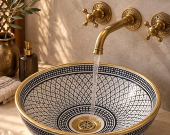 Può includere: Un lavabo da appoggio rotondo in ceramica con motivo geometrico bianco e nero, bordo e scarico dorati. Un rubinetto a parete in ottone con dettagli circolari complessi eroga acqua nel bacino, posizionato su un piano d'appoggio in pietra chiara.