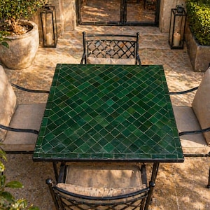 May include: Outdoor dining set featuring a square table with a green tiled top and black metal frame. Four chairs with patterned backs and beige cushions surround the table. The setting is an outdoor patio with stone flooring and potted plants.