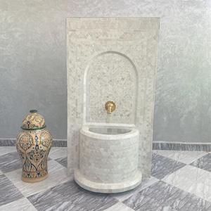 Puede incluir: Fuente de pared decorativa de mosaico blanco con grifo de latón. A la izquierda hay un jarrón de cerámica estampado con tapa. La fuente tiene una pila curva y una alcoba arqueada. El suelo está embaldosado con un patrón de tablero de ajedrez gris y blanco.