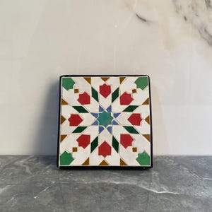 May include: Square decorative tile with a geometric starburst pattern. The design features red, green, blue, and gold accents on a white background, framed by a black border. The tile is likely ceramic or similar material.