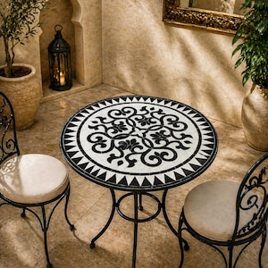 May include: A small outdoor bistro set with a round mosaic tile table and two wrought iron chairs. The table features a black and white floral design. The chairs have cream-colored cushions. The set is ideal for a patio or balcony.