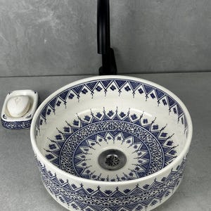 Op de afbeelding: Een witte keramische wastafel met een blauw geometrisch patroon. De ronde wastafel heeft een decoratief ontwerp rond de rand en in de kom. Een zwarte kraan is achter de wastafel gemonteerd en een bijpassende zeepbakje staat links.