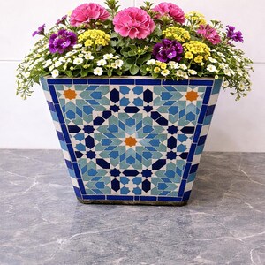 Op de afbeelding: Een vierkante mozaïek bloempot gevuld met een verscheidenheid aan kleurrijke bloemen. De bloempot heeft een geometrisch patroon in blauwtinten, met oranje accenten. De bloemen omvatten roze, paarse, gele en witte bloemen.