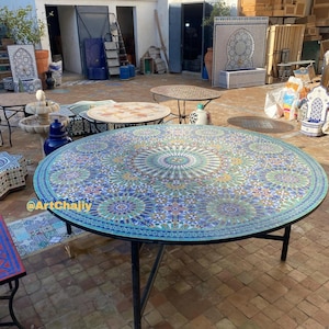 May include: A large, round mosaic tile table with a black metal frame. The tabletop features a detailed pattern in shades of blue, green, and gold. The table is outdoors on a brick patio. The watermark "@ArtChajiy" is visible.