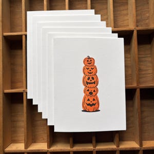 May include: A stack of white greeting cards featuring a centered illustration of five orange pumpkins stacked vertically. Each pumpkin has a unique black carved jack-o'-lantern face, ranging from simple circular eyes to jagged smiles, arranged in a column.