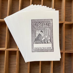 May include: A stack of white paper prints featuring a black and white illustration of a person reading in a window, with the text "OWNETH THIS BOOK" below the image. The prints are stacked on a wooden surface.