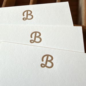 May include: Three rectangular white note cards stacked in a staggered arrangement. Each card features a centered, embossed letter B in a metallic gold script font. The paper has a visible textured finish, and the cards are clean and blank.