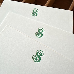 May include: Three rectangular white note cards stacked in a staggered arrangement. Each card features a centered, embossed letter S in a dark green, ornate, calligraphic font with a subtle shadow effect on the paper surface.