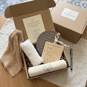 May include: A cardboard box containing a pet comfort kit with a tan towel, a white plush toy, a grey circular lick mat, a wooden care paddle with text, and a rolled cream cloth with a dog illustration. A black marker and a closed box are nearby.