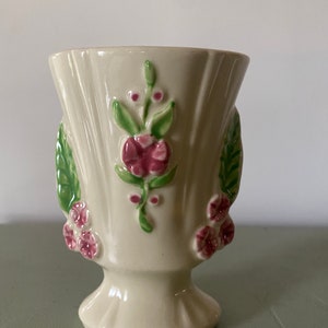 May include: A white ceramic vase with a pedestal base. The vase is decorated with raised pink and green flowers.