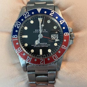 May include: A stainless steel Rolex Oyster Perpetual Submariner watch with a black face, red and blue bezel, and a date window at the 3 o'clock position. The watch has a silver-colored band and a silver-colored crown.