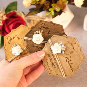 May include: Wooden wedding favor tags with floral accents. Each tag features the names "Jason & Rebecca" and the date "10.06.2026". The tags are tied with twine and adorned with white flowers.