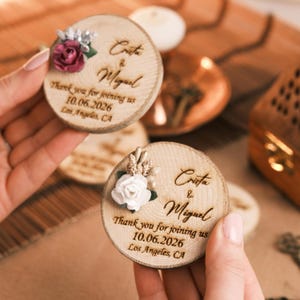 May include: Wooden round favors with floral accents. The favors are engraved with the names "Crista & Miguel", the date "10.06.2026", and the location "Los Angeles, CA". One favor has a burgundy rose, and the other has a white rose.