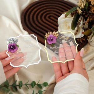 May include: Two clear acrylic wedding favors with floral accents. One favor has a silver border and the other has a gold border. Each favor has a purple rose and text that reads "Thank you for joining us".