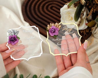 Puede incluir: Dos recuerdos de boda de acrílico transparente con detalles florales. Uno tiene un borde plateado y el otro un borde dorado. Cada recuerdo tiene una rosa morada y el texto "Gracias por acompañarnos".