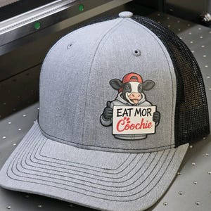 May include: Gray trucker hat with a black mesh back. The hat features a cartoon cow wearing a red cap and hoodie, holding a sign that reads "EAT MOR Coochie".