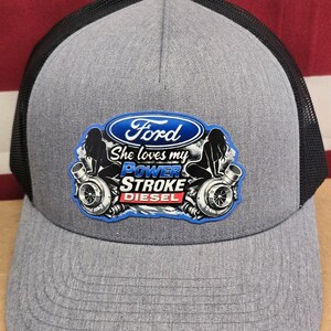 May include: Gray trucker hat with a black mesh back. The front features a Ford logo and the text "She loves my Power Stroke Diesel" with a graphic of a woman and a turbocharger.