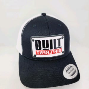 May include: Black and white trucker hat with a white mesh back. The hat has a black and white rectangular patch that says "BUILT DIFFERENT" in bold white letters with the word "DIFFERENT" in red.