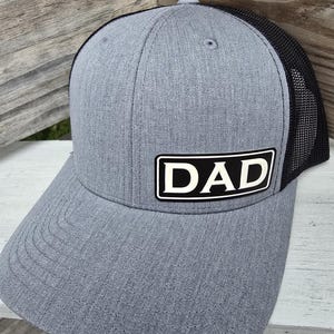May include: Gray trucker hat with a black mesh back and a curved brim. The hat features a rectangular patch with the word "DAD" in white block letters on a black background. The hat is made of a textured fabric.