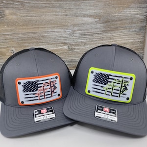 May include: Two gray trucker hats with black mesh back panels. Both hats feature a white American flag with a red, orange, or yellow outline. The flag has a silhouette of a lineman working on power lines. The hats are labeled "Richardson 112".