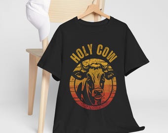 May include: Black t-shirt with a distressed graphic of a cow's head inside a sunset-colored circle. The words "HOLY COW" are arched above the cow. The shirt is hanging on a wooden hanger.