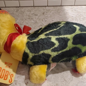 Könnte beinhalten: Ein Plüsch-Schildkrötenspielzeug mit gelbem Kopf und Gliedmaßen, einem schwarz-grün gemusterten Panzer und einem roten Band um den Hals. Sie trägt einen kleinen orangefarbenen Hut und hat ein gelbes Etikett mit der Aufschrift WON AT BOB'S.