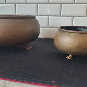 Puede incluir: Dos cuencos antiguos de bronce con cuerpos redondeados y patas de garra. Los cuencos tienen una textura martillada y una pátina oscura. Uno de los cuencos es más grande que el otro. Se encuentran sobre una alfombra negra con borde rojo.