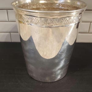 Puede incluir: Una cubitera de metal plateado con un patrón decorativo de flores y vides alrededor del borde. La cubitera tiene forma cilíndrica y una superficie muy reflectante.