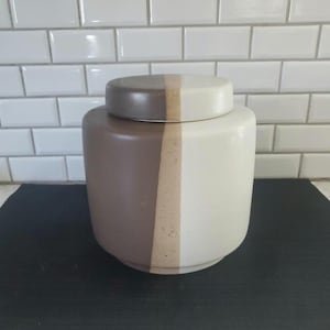 Pode incluir: Um pote de cerâmica com tampa, com um design em blocos de cores. O pote é dividido em três secções verticais: castanho, bege e branco sujo. A tampa espelha as secções castanhas e branco sujo. O pote é cilíndrico.