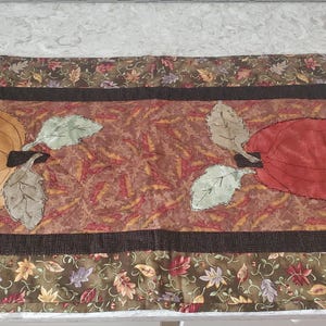 Peut inclure: Un chemin de table rectangulaire en tissu avec un panneau central à motif de feuilles marron, rouges et orange. Le chemin présente deux grandes formes de fruits appliquées, une jaune et une rouge, ornées de feuilles en tissu vert et gris.