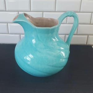May include: A turquoise ceramic pitcher with a curved handle and a spout. The interior is a light brown color, contrasting with the exterior's glossy, swirling design. The pitcher is a decorative item.